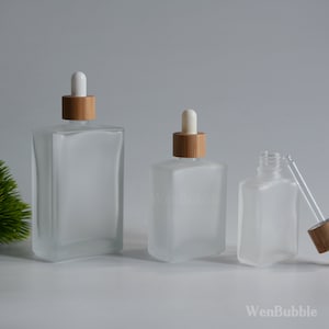 Puede incluir: Tres botellas de vidrio esmerilado con tapas de bambú y goteros blancos. Las botellas son de diferentes tamaños y están dispuestas en fila sobre una superficie blanca.