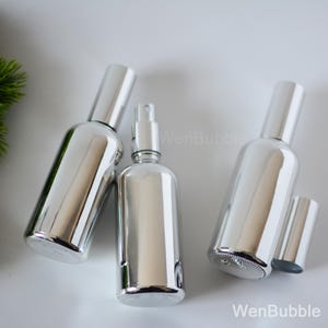 May include: Three silver glass bottles with spray tops. The bottles are empty and have a reflective surface.