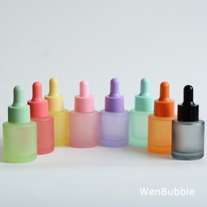 May include: A collection of frosted glass dropper bottles in various pastel colors, including green, pink, yellow, purple, and orange. Each bottle has a matching colored dropper top. The bottles are arranged in a row against a white background.