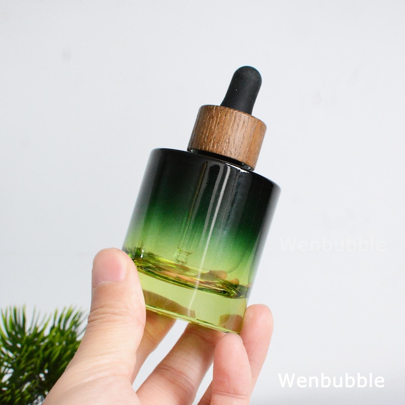 May include: A green-tinted glass dropper bottle with a wooden cap and black dropper. The bottle has a gradient effect, transitioning from green to clear. The bottle is held in a hand.
