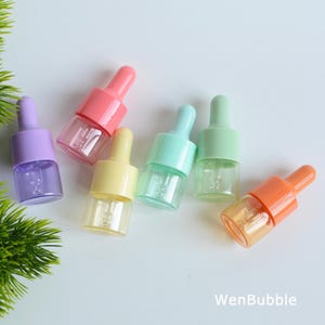 Może przedstawiać: Kolekcja małych, kolorowych butelek z zakraplaczem. Butelki mają przezroczyste szklane korpusy i pastelowe nakrętki w odcieniach różu, fioletu, żółci, miętowej zieleni i pomarańczy. Butelki ułożone są na białej powierzchni.