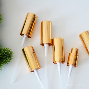 Könnte beinhalten: Mehrere goldfarbene Sprühflaschenaufsätze mit weißen Stielen sind vor einem weißen Hintergrund angeordnet. Die Aufsätze sind zylindrisch und haben eine glänzende, metallische Oberfläche. Die Stiele sind dünn und transparent.