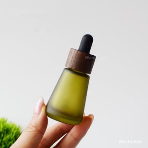 May include: A frosted olive green glass essential oil bottle with a wooden cap and black dropper. The bottle has a conical shape and is held by a hand against a soft white background. The wooden cap has a dark brown color.