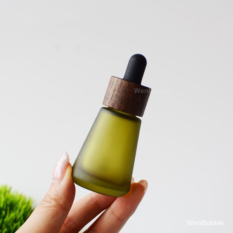 May include: A frosted olive green glass essential oil bottle with a wooden cap and black dropper. The bottle has a conical shape and is held by a hand against a soft white background. The wooden cap has a dark brown color.
