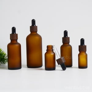 Puede incluir: Un conjunto de cinco frascos cuentagotas de vidrio esmerilado color ámbar de varios tamaños. Cada frasco tiene un cuello con patrón de vetas de madera marrón oscuro y una perilla de goma negra. Un frasco está abierto con la pipeta fuera.