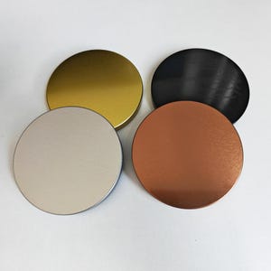 May include: Four circular metal lids in gold, black, copper, and silver. The lids are arranged in a circular pattern, each with a smooth, reflective surface. The gold lid is at the top left, the black lid is at the top right, the copper lid is at the bottom right, and the silver lid is at the bottom left.