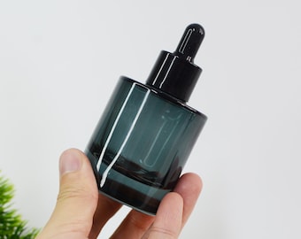 May include: A dark teal glass dropper bottle with a black dropper top. The bottle has a cylindrical shape and is held in a hand. The bottle is empty and ready to be filled with essential oils or other liquids.
