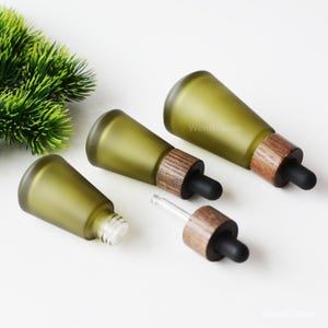 May include: Four olive green glass dropper bottles with wooden collars and black rubber tips. One bottle has a clear glass dropper inserted. The bottles are arranged on a white surface. The text "WenBubble" is visible on the bottles.