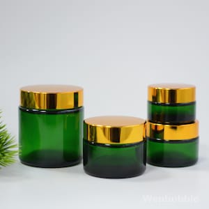 Peut inclure: Quatre pots en verre vert avec des couvercles dorés. Les pots sont cylindriques et de tailles différentes, avec un pot empilé sur deux autres. Les pots sont vides et prêts à être remplis.