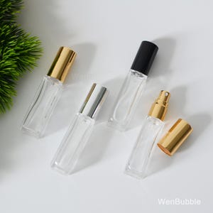 Könnte beinhalten: Vier transparente Parfümflaschen mit verschiedenfarbigen Kappen: Gold, Silber und Schwarz. Eine Flasche hat einen goldenen Sprühkopf. Die rechteckigen Flaschen sind leer und bereit zum Befüllen. Der Hintergrund ist weiß.