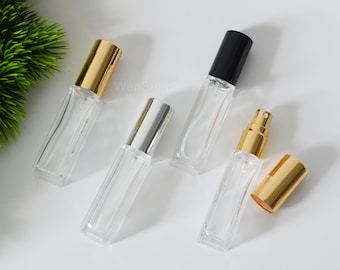 Op de afbeelding: Vier heldere glazen parfumflesjes met verschillend gekleurde doppen: goud, zilver en zwart. Eén flesje heeft een gouden sproeikop. De rechthoekige flesjes zijn leeg en klaar om te worden gevuld. De achtergrond is wit.