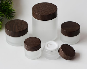 May include: A collection of frosted glass jars with dark brown wooden lids. The jars vary in size, with one jar open, revealing a white interior. The jars are arranged on a white surface. The text "WenBubble" is visible.
