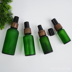 Puede incluir: Cuatro frascos pulverizadores de vidrio esmerilado verde de varios tamaños, cada uno con un cuello de madera oscura y una boquilla de plástico negro. Un frasco tiene su tapa negra retirada al lado. El texto wenbubble es visible.