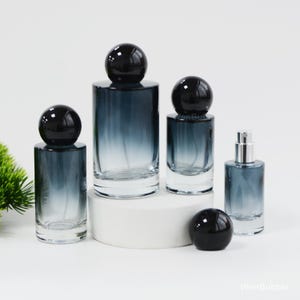 May include: A collection of glass perfume bottles in various sizes, featuring a gradient blue-gray color. Each bottle has a glossy black spherical cap. The bottles are arranged on a white surface, with a small green plant in the background.