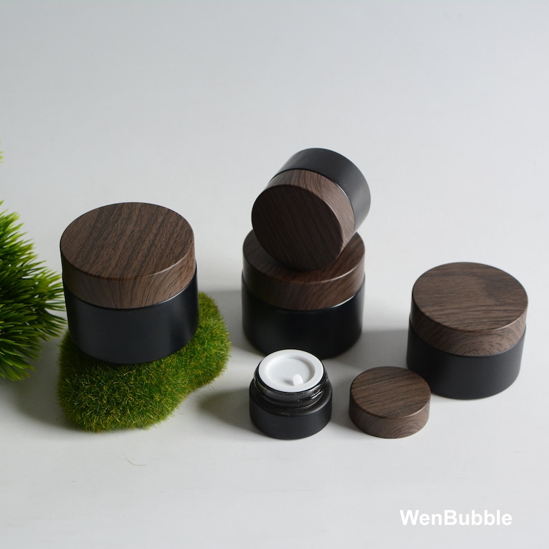 May include: Five black glass jars with wooden lids. The jars are stacked and arranged on a white surface with green moss.
