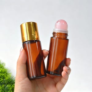 May include: Two amber glass roller bottles. One bottle features a shiny gold cap, while the other is open, revealing a smooth, light pink roller ball applicator at the top. Both containers are cylindrical and made of translucent brown glass.