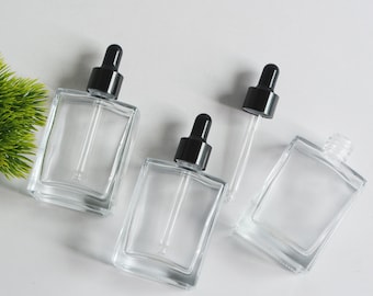 May include: Three clear glass bottles with black dropper tops. The bottles are square shaped and have a clear glass dropper.