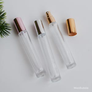 Puede incluir: Tres frascos de perfume de vidrio transparente con tapas metálicas en oro rosa, plata y oro. Un frasco tiene la tapa quitada. Los frascos están vacíos y listos para ser llenados. Los frascos están sobre una superficie blanca.