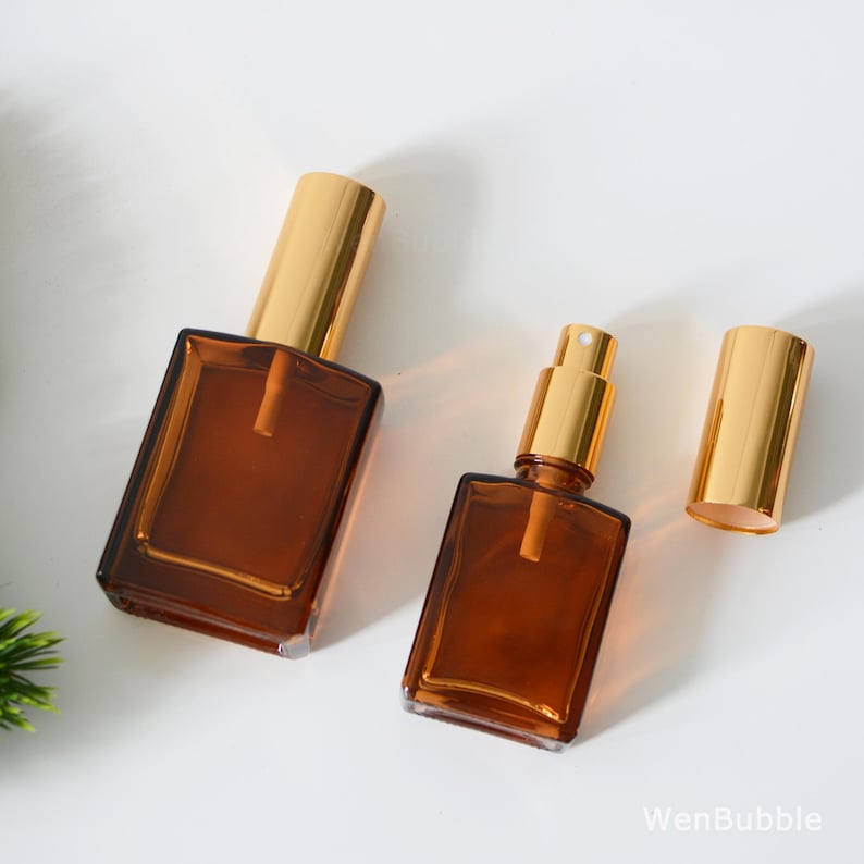 May include: Two amber glass bottles with gold spray tops and a separate gold spray top. The bottles are square shaped and have a frosted glass look.