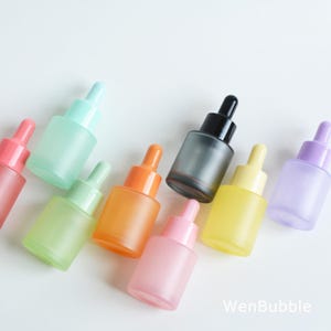 May include: A collection of frosted glass dropper bottles in various pastel colors, including pink, green, orange, yellow, and purple. Each bottle has a matching colored dropper top and a clear glass body. The bottles are arranged on a white surface.