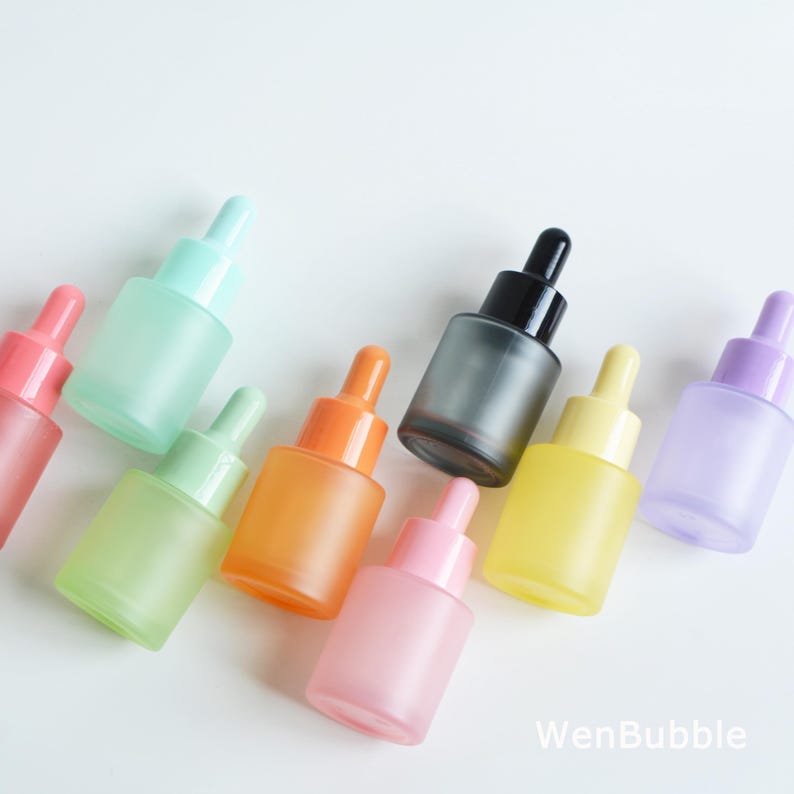 May include: A collection of frosted glass dropper bottles in various pastel colors, including pink, green, orange, yellow, and purple. Each bottle has a matching colored dropper top and a clear glass body. The bottles are arranged on a white surface.