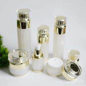 Könnte beinhalten: Eine Sammlung weiß-goldener Kosmetikbehälter, darunter Flaschen und Tiegel. Die Behälter haben eine gerippte Textur und goldfarbene Akzente auf den Deckeln und Böden. Das Bild zeigt eine Vielzahl von Größen und Formen.