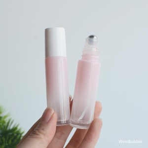 May include: Two pink and white roll-on bottles. One bottle has a white cap, and the other has a silver rollerball applicator. The bottles are held in a hand against a white background. The bottles have a gradient pink color.