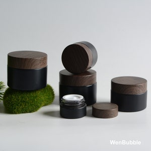 May include: Five black glass jars with wooden lids. The jars are different sizes and are arranged in a pyramid shape. The jars are on a white surface with a green moss-like material in the background.