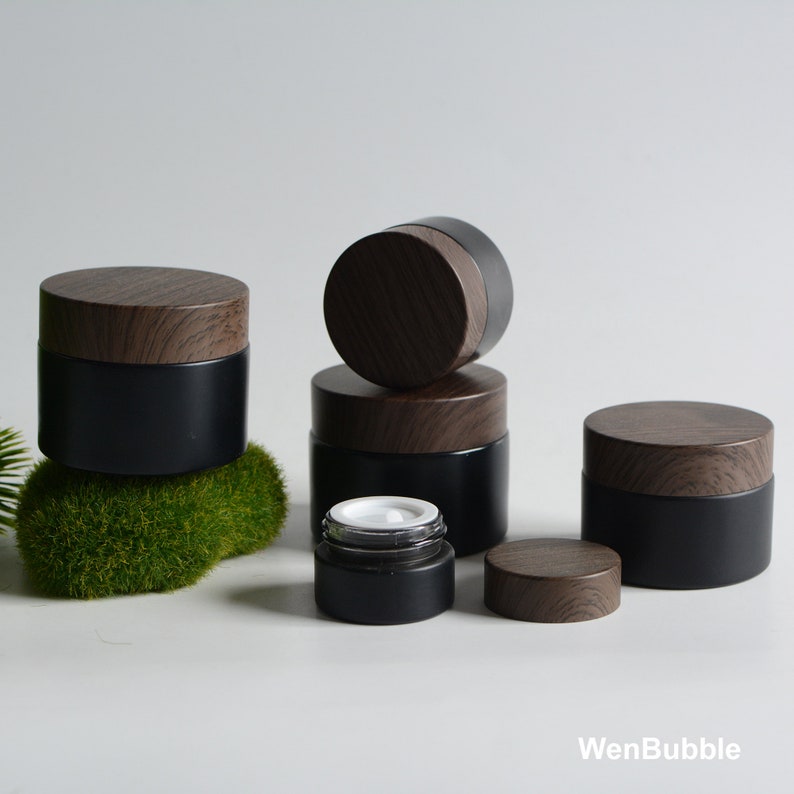 May include: Five black glass jars with wooden lids. The jars are different sizes and are arranged in a pyramid shape. The jars are on a white surface with a green moss-like material in the background.