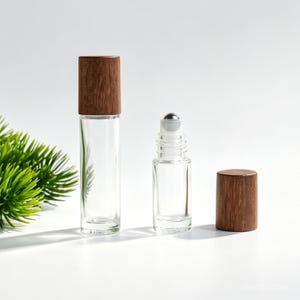 Könnte beinhalten: Zwei durchsichtige Glas-Roll-on-Fläschchen mit dunkelbraunen Holzmaserungsverschlüssen. Eine Flasche ist verschlossen, während die andere offen ist und eine silberne Metallkugel an der Spitze zeigt. Die Flaschen sind zylindrisch und glatt.