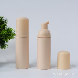 May include: Three beige foam pump bottles, one with the pump head open, and one with the cap off. The bottles are cylindrical with rounded tops. The background is white with a green plant in the upper left corner. The text "WenBubble" is in the lower right corner.