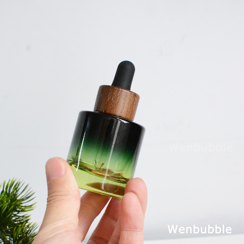 May include: A green and yellow glass dropper bottle with a wooden cap and black dropper. The bottle is held in a hand. The bottle has a gradient color, transitioning from green to yellow. The bottle is likely for essential oils or serums.