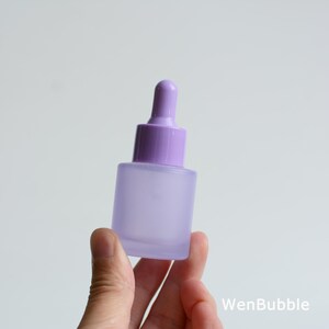 May include: A small, frosted glass dropper bottle with a square base and a purple dropper top. The bottle is held in a hand against a white background. The bottle is a soft lavender color.