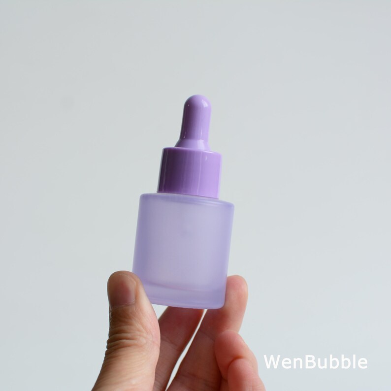 May include: A small, frosted glass dropper bottle with a square base and a purple dropper top. The bottle is held in a hand against a white background. The bottle is a soft lavender color.