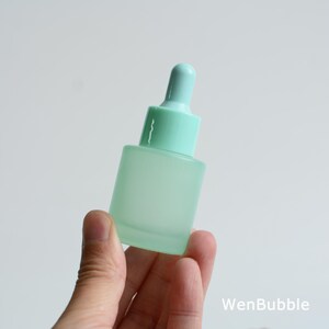 May include: A frosted glass dropper bottle in a soft mint green color. The bottle has a matching mint green dropper top. The bottle is held in a hand against a white background. The text "WenBubble" is in the bottom right corner.