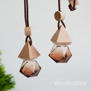 May include: Two hanging car air fresheners. Each has a geometric glass bottle in shades of brown, topped with a wooden pyramid and a wooden cylinder. The air fresheners are suspended by brown cords. The background is a soft, neutral color.