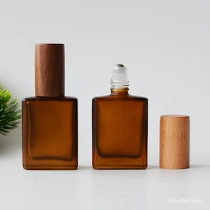 May include: Three amber glass bottles with wooden caps. One bottle has a roll-on applicator, and the other has a wooden cap. The bottles are rectangular and sit on a white surface. The wooden caps are cylindrical.