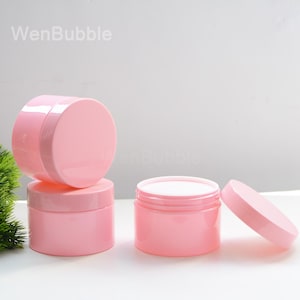 Puede incluir: Frascos cosméticos rosas con tapas, uno de los frascos está abierto, revelando un interior blanco. Los frascos están apilados y dispuestos sobre una superficie blanca. El texto "WenBubble" es visible.