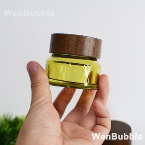 May include: A small, clear glass jar with a yellow tint, topped with a dark brown wooden lid. The jar is held in a hand against a white background. The text "WenBubble" is visible in the upper left and lower right corners.