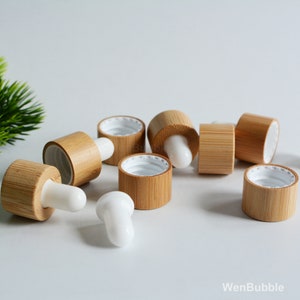 May include: A group of eight wooden bottle caps with white droppers. The caps are made of natural bamboo and have a light brown color. The droppers are made of white plastic and are designed for essential oils or other liquids.