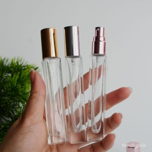 Puede incluir: Tres frascos de perfume de vidrio transparente con atomizadores metálicos dorados, plateados y oro rosa. Los frascos son rectangulares con un diseño acanalado y se sostienen en una mano.