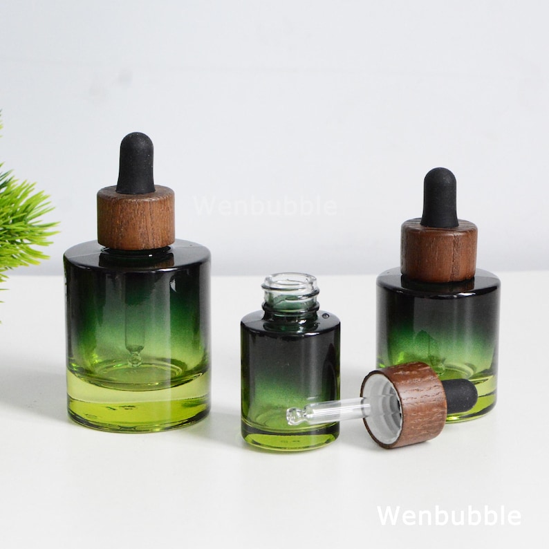 May include: Three green glass dropper bottles with wooden caps. The bottles have a gradient color, transitioning from a dark green at the top to a lighter green at the bottom. One bottle has its dropper exposed. The bottles are on a white surface.