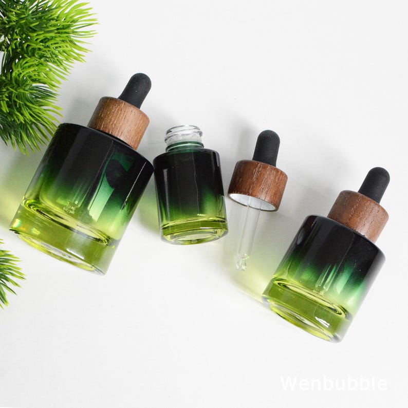 May include: Four glass dropper bottles with a gradient green color, wooden caps, and black droppers. The bottles are arranged on a white surface, with a sprig of green foliage in the upper left corner. The bottles vary in size.