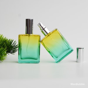 May include: Two rectangular glass perfume bottles with a yellow to green gradient. One bottle is upright, the other is tilted, and a silver cap is next to it. The bottles have silver sprayers.