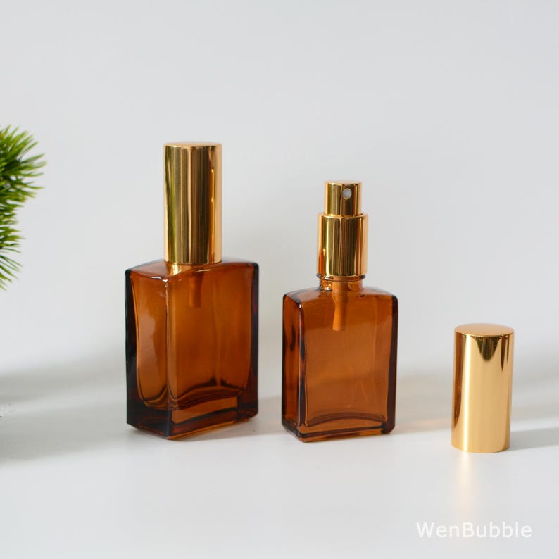 May include: Two amber glass bottles with gold spray tops and a separate gold spray top. The bottles are square shaped with rounded edges.