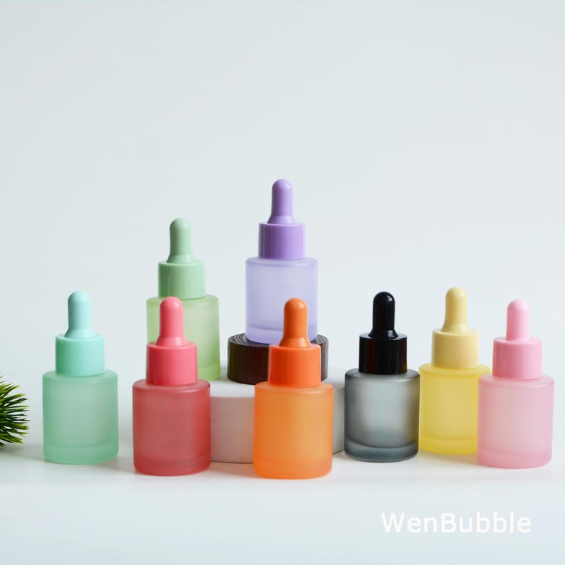 May include: A collection of frosted glass dropper bottles in various colors, including green, pink, orange, and purple. Each bottle has a matching colored dropper top. The bottles are arranged on a white surface.