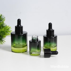 Puede incluir: Tres frascos cuentagotas de vidrio verde-negro de varios tamaños. Una botella está abierta y otra tiene un cuentagotas al lado. Las botellas tienen un efecto de color degradado, que va del verde claro en la base al negro oscuro en la parte superior.