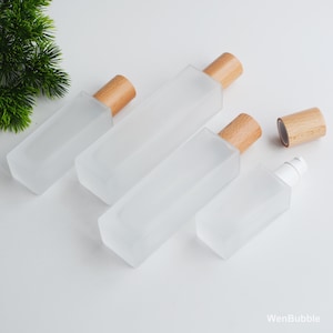 Pode incluir: Quatro frascos de vidro fosco com tampas de madeira dispostos sobre uma superfície branca. Os frascos variam em tamanho e formato, com um design retangular. Uma tampa está separada. Os frascos são adequados para armazenar líquidos.