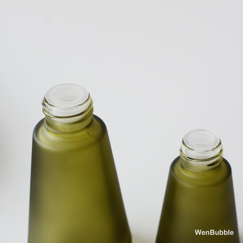 May include: Two olive green glass bottles with white tops. The bottles have a matte finish and a conical shape. The tops are clear and have a threaded design. The bottles are on a white background. The text "WenBubble" is visible on the bottom right.