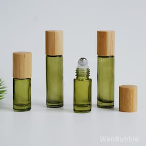 May include: Four cylindrical olive green glass roller bottles with light brown wooden caps. Three bottles are capped, while one central bottle is uncapped, revealing a silver metal roller ball. The text WenBubble is visible in the bottom right corner.